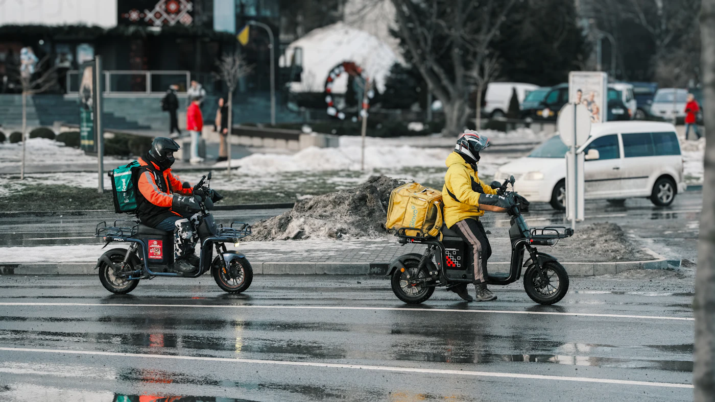 Dois entregadores em scooters numa rua molhada de chuva
